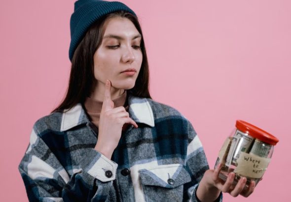 Pensive woman with a savings jar labeled 'where to next' on a pink background.