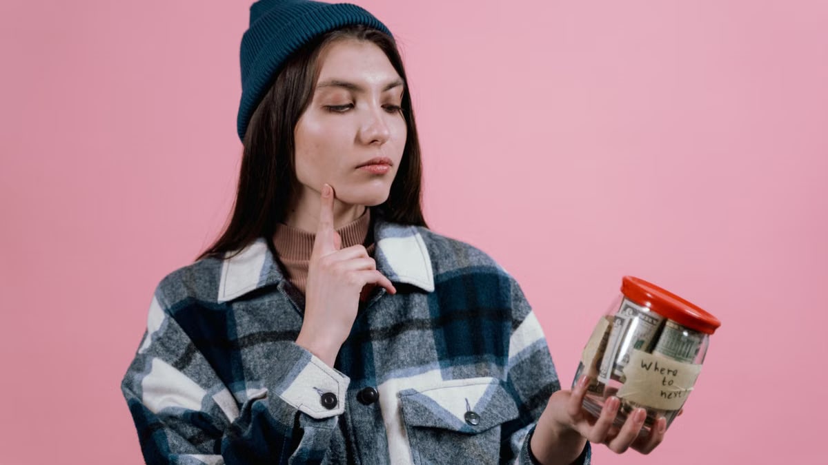 Pensive woman with a savings jar labeled 'where to next' on a pink background.